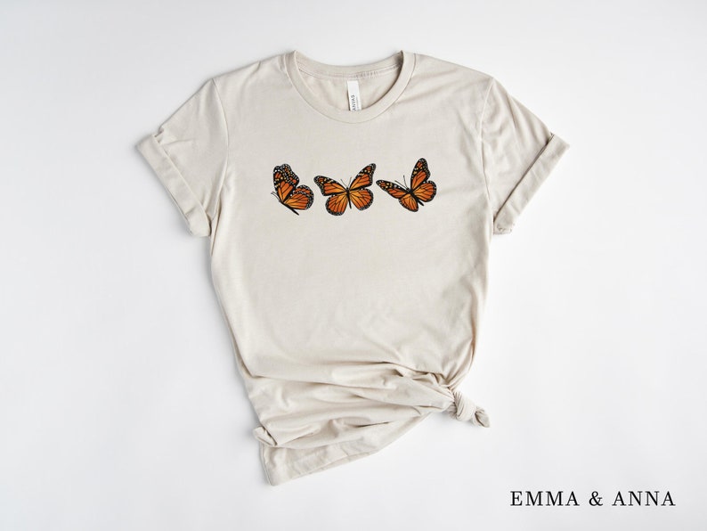 Może przedstawiać: Jasny beżowy t-shirt z trzema ilustracjami pomarańczowo-czarnych motyli monarchy. Motyle ułożone są poziomo na piersi. Tekst "EMMA & ANNA" jest wydrukowany na dole po prawej stronie.