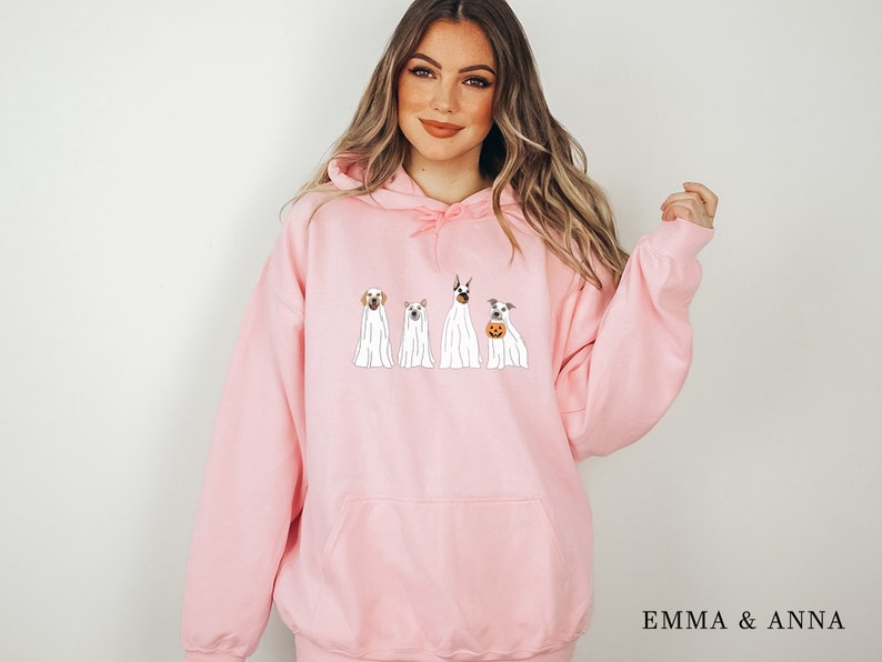 May include: Pink hooded sweatshirt with a graphic of four dogs dressed as ghosts for Halloween. The text "EMMA & ANNA" is printed on the bottom right corner of the sweatshirt.