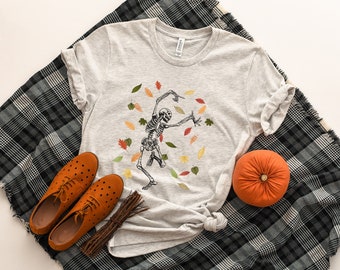Puede incluir: Una camiseta gris con un esqueleto bailando en un montón de hojas de otoño. La camiseta está colocada sobre una manta de cuadros negros y blancos con una pequeña calabaza naranja.