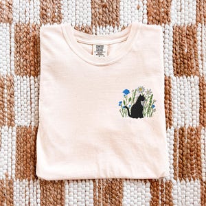 May include: A light peach-colored t-shirt with a crew neck. The shirt features a black cat silhouette embroidered among blue and white flowers. The shirt is folded neatly on a woven, checkered rug.