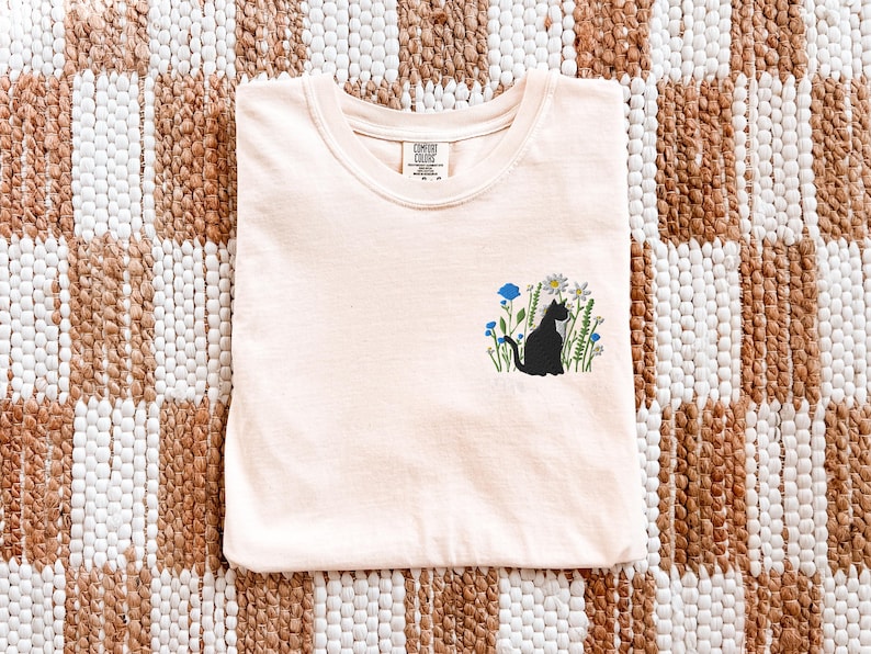 May include: A light peach-colored t-shirt with a crew neck. The shirt features a black cat silhouette embroidered among blue and white flowers. The shirt is folded neatly on a woven, checkered rug.