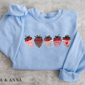 Puede incluir: Una sudadera azul claro con un gr&aacute;fico de cinco fresas cubiertas de chocolate. Cada fresa tiene un dise&ntilde;o diferente, que incluye rayas blancas, remolinos de chocolate y un coraz&oacute;n rojo.