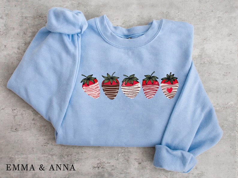Puede incluir: Una sudadera azul claro con un gr&aacute;fico de cinco fresas cubiertas de chocolate. Cada fresa tiene un dise&ntilde;o diferente, que incluye rayas blancas, remolinos de chocolate y un coraz&oacute;n rojo.