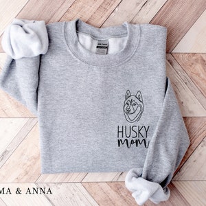 Pode incluir: Camisola cinzenta com um contorno preto de um c&atilde;o husky e o texto "Husky Mom" na frente.