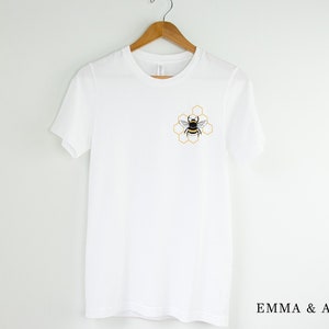 May include: White t-shirt with a bee and honeycomb design on the front. The text "EMMA & ANNA" is printed on the bottom of the shirt.