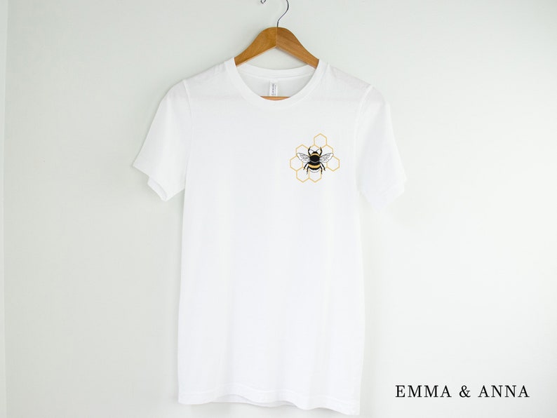 May include: White t-shirt with a bee and honeycomb design on the front. The text "EMMA & ANNA" is printed on the bottom of the shirt.