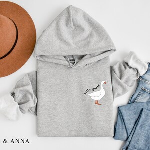 K&ouml;nnte beinhalten: Ein grauer Kapuzenpullover mit einer wei&szlig;en G&auml;nse-Illustration und dem Text "silly goose" auf der Vorderseite.