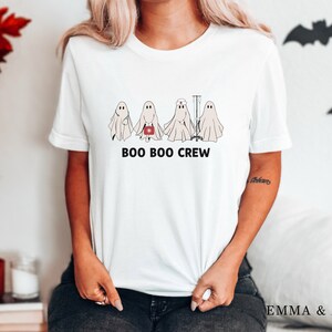 Puede incluir: Camiseta blanca con un gr&aacute;fico de cinco fantasmas. Los fantasmas llevan gorros de enfermera y llevan suministros m&eacute;dicos. El texto "BOO BOO CREW" est&aacute; impreso debajo de los fantasmas.