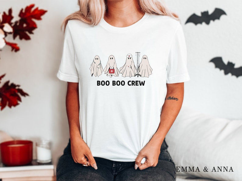 Puede incluir: Camiseta blanca con un gr&aacute;fico de cinco fantasmas. Los fantasmas llevan gorros de enfermera y llevan suministros m&eacute;dicos. El texto "BOO BOO CREW" est&aacute; impreso debajo de los fantasmas.