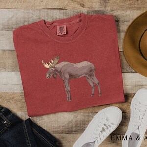 May include: A red t-shirt with a brown moose graphic. The shirt is laid out on a wooden surface with a brown hat, blue jeans, and white sneakers.