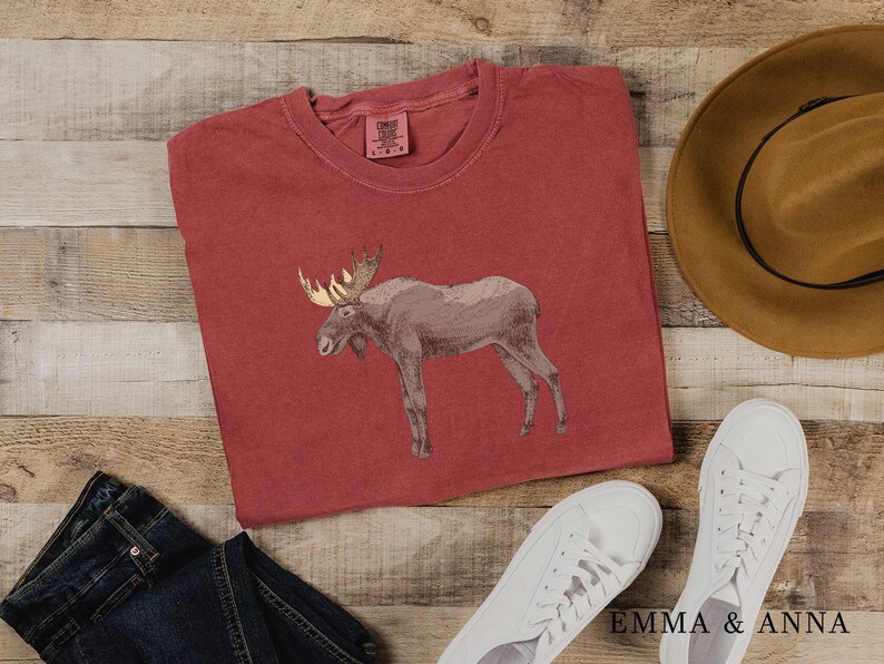 May include: A red t-shirt with a brown moose graphic. The shirt is laid out on a wooden surface with a brown hat, blue jeans, and white sneakers.