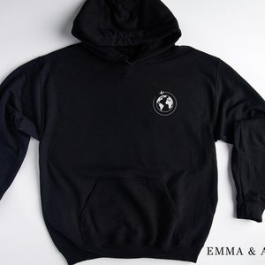 May include: Black hooded sweatshirt with a white graphic of a globe with a plane flying above it. The text "EMMA & ANNA" is printed on the bottom right corner of the image.