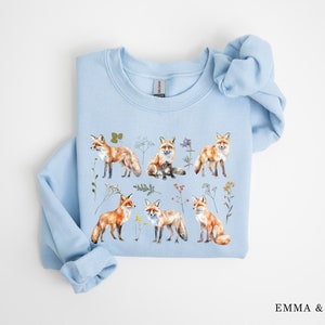 May include: A light blue sweatshirt with a watercolor print of foxes and wildflowers. The sweatshirt features a variety of foxes in different poses, surrounded by delicate wildflowers in shades of pink, purple, and yellow.
