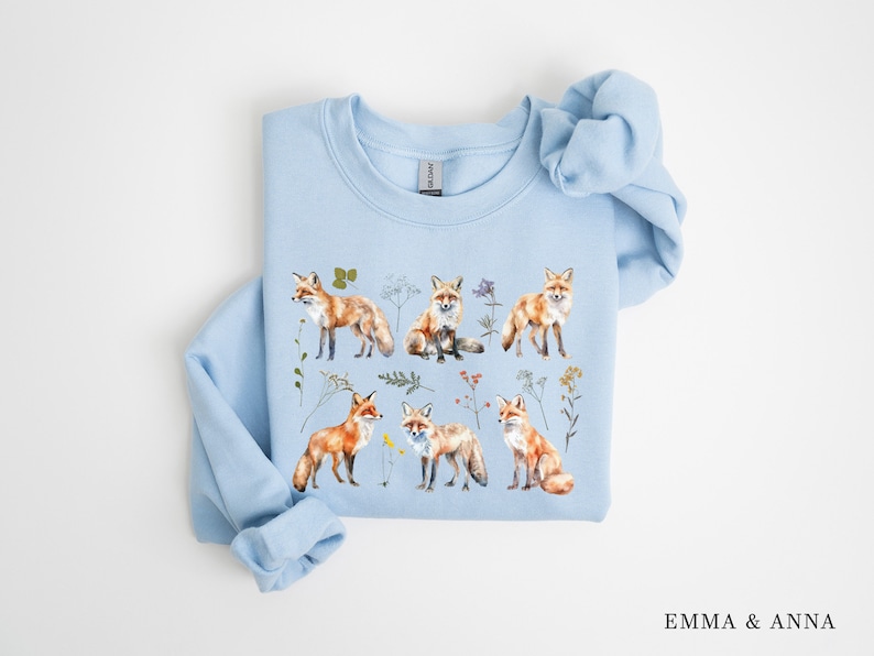 May include: A light blue sweatshirt with a watercolor print of foxes and wildflowers. The sweatshirt features a variety of foxes in different poses, surrounded by delicate wildflowers in shades of pink, purple, and yellow.