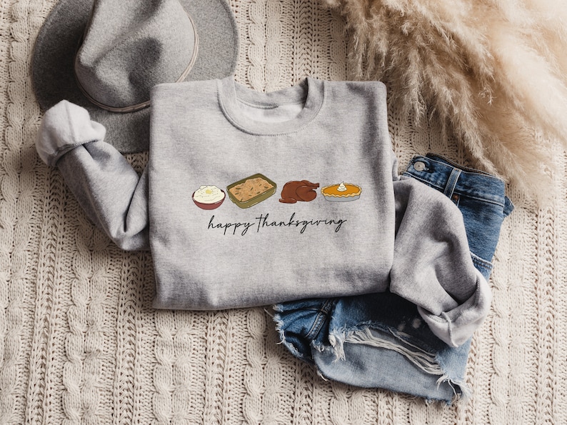 K&ouml;nnte beinhalten: Ein graues Sweatshirt mit einem Thanksgiving-Motiv, das einen Truthahn, Kartoffelp&uuml;ree, F&uuml;llung und K&uuml;rbiskuchen zeigt. Der Text "happy Thanksgiving" ist unter den Speisen gedruckt.