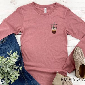 Peut inclure: Un t-shirt &agrave; manches longues de couleur mauve avec un motif repr&eacute;sentant une boisson au caf&eacute; sur la poitrine gauche. Le t-shirt est pos&eacute; sur une surface en bois blanc avec un jean bleu, une plante en pot et deux paires de bottines beige.