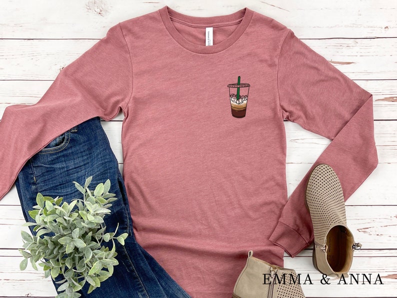 Peut inclure: Un t-shirt &agrave; manches longues de couleur mauve avec un motif repr&eacute;sentant une boisson au caf&eacute; sur la poitrine gauche. Le t-shirt est pos&eacute; sur une surface en bois blanc avec un jean bleu, une plante en pot et deux paires de bottines beige.