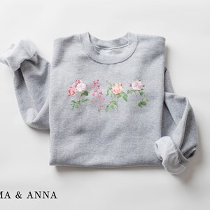 May include: Gray sweatshirt with a floral print of pink and white roses on the front. The text "EMMA & ANNA" is printed below the image.