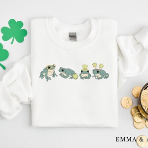 Puede incluir: Sudadera blanca con un tr&eacute;bol verde y cuatro ranas verdes que llevan sombreros y gafas. Las ranas llevan un sombrero verde con un tr&eacute;bol, un sombrero verde con un sombrero de copa blanco, un sombrero verde con un sombrero de copa blanco y un sombrero verde con un tr&eacute;bol.