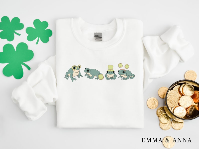 Puede incluir: Sudadera blanca con un tr&eacute;bol verde y cuatro ranas verdes que llevan sombreros y gafas. Las ranas llevan un sombrero verde con un tr&eacute;bol, un sombrero verde con un sombrero de copa blanco, un sombrero verde con un sombrero de copa blanco y un sombrero verde con un tr&eacute;bol.