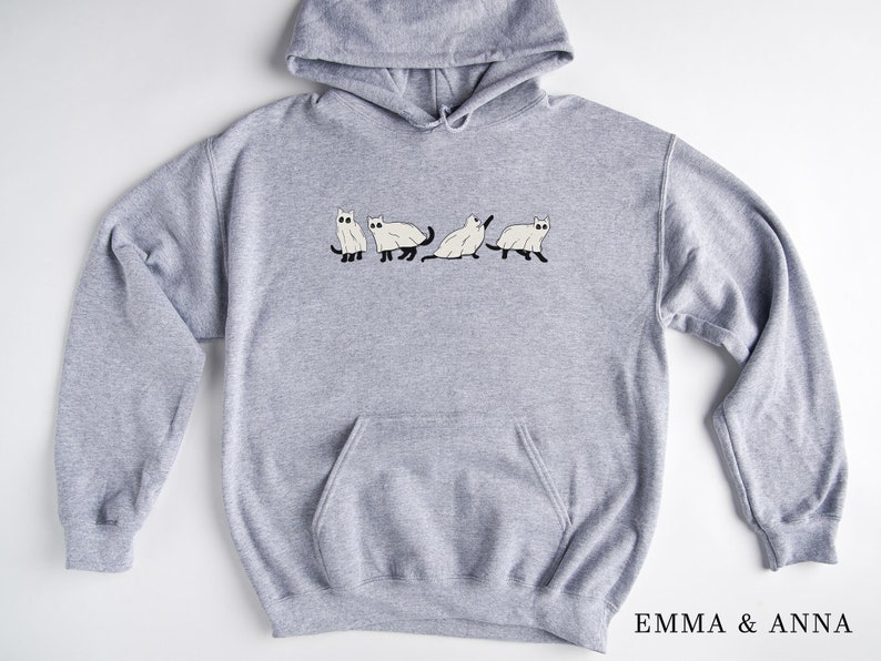 Puede incluir: Una sudadera con capucha gris con un gr&aacute;fico de cuatro gatos blancos en la parte delantera. Los gatos est&aacute;n en l&iacute;nea, y el del medio est&aacute; sentado. El texto "EMMA & ANNA" est&aacute; impreso en la esquina inferior derecha de la sudadera.