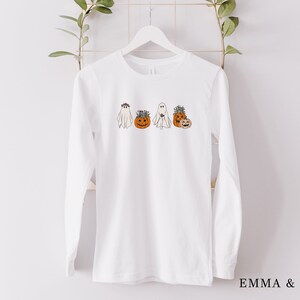 May include: White long-sleeve t-shirt with a graphic of four ghosts and pumpkins. The ghosts are wearing flower crowns and the pumpkins have faces.