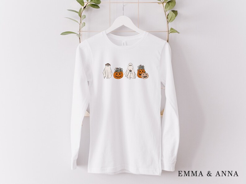 May include: White long-sleeve t-shirt with a graphic of four ghosts and pumpkins. The ghosts are wearing flower crowns and the pumpkins have faces.