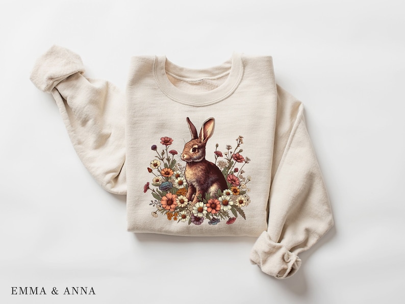 Pu&ograve; includere: Felpa color crema con un coniglio marrone circondato da fiori colorati. Il coniglio guarda a destra. I fiori includono tonalit&agrave; di rosso, arancione e giallo. Il testo "EMMA & ANNA" &egrave; in basso. Capo d'abbigliamento comodo.