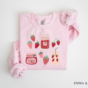May include: Pink sweatshirt with a strawberry jam and strawberry drink graphic. The graphic features jars of strawberry jam and a glass of strawberry juice with a straw. There are also strawberries scattered around the graphic.