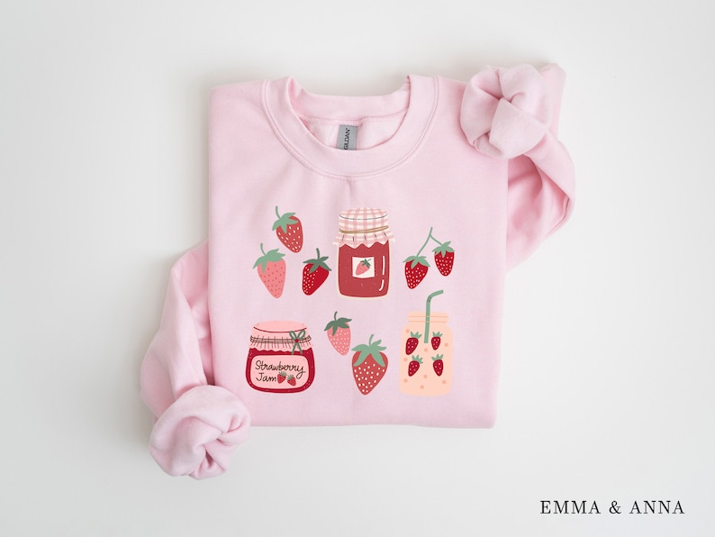May include: Pink sweatshirt with a strawberry jam and strawberry drink graphic. The graphic features jars of strawberry jam and a glass of strawberry juice with a straw. There are also strawberries scattered around the graphic.