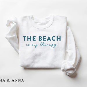 以下が含まれることがあります： ターコイズブルーの文字で「THE BEACH is my therapy」と書かれた白いスウェットシャツです。