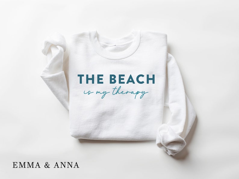 以下が含まれることがあります： ターコイズブルーの文字で「THE BEACH is my therapy」と書かれた白いスウェットシャツです。