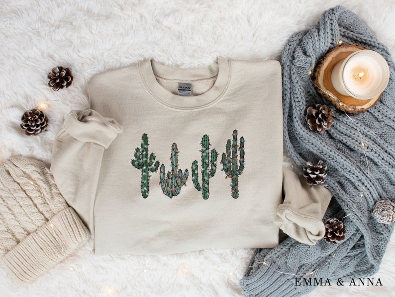 May include: A beige sweatshirt with a graphic of four cacti with green leaves and red lights on them. The sweatshirt is on a white fuzzy surface with pine cones and string lights.