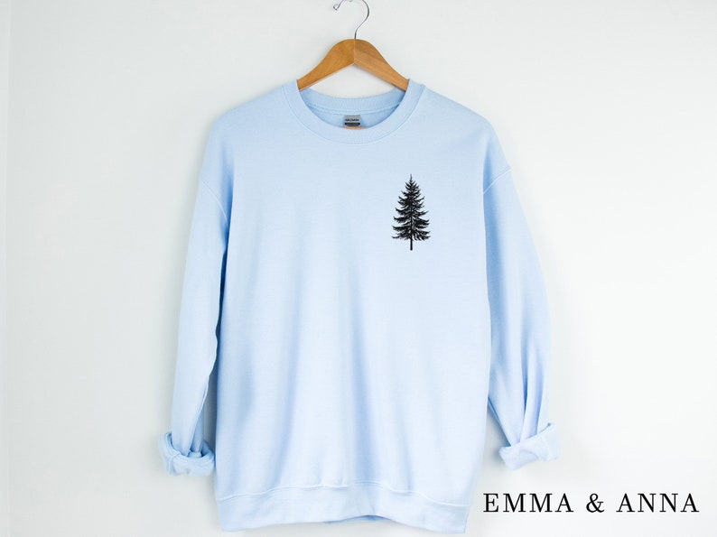 May include: A light blue sweatshirt with a black silhouette of a pine tree on the front. The sweatshirt is hanging on a wooden hanger. The text "EMMA & ANNA" is printed on the bottom right corner of the image.