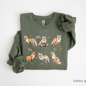 May include: A green sweatshirt with a print of multiple orange foxes and floral details. The sweatshirt has a crew neck and long sleeves.