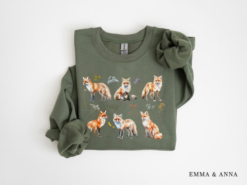 May include: A green sweatshirt with a print of multiple orange foxes and floral details. The sweatshirt has a crew neck and long sleeves.