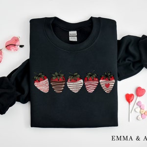Puede incluir: Sudadera negra con un gr&aacute;fico de cinco fresas ba&ntilde;adas en chocolate y chocolate blanco. Las fresas tienen corazones rojos en el centro. La sudadera tiene cuello redondo y mangas largas.
