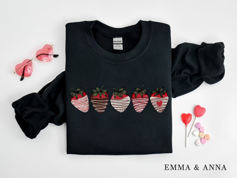 Puede incluir: Sudadera negra con un gr&aacute;fico de cinco fresas ba&ntilde;adas en chocolate y chocolate blanco. Las fresas tienen corazones rojos en el centro. La sudadera tiene cuello redondo y mangas largas.