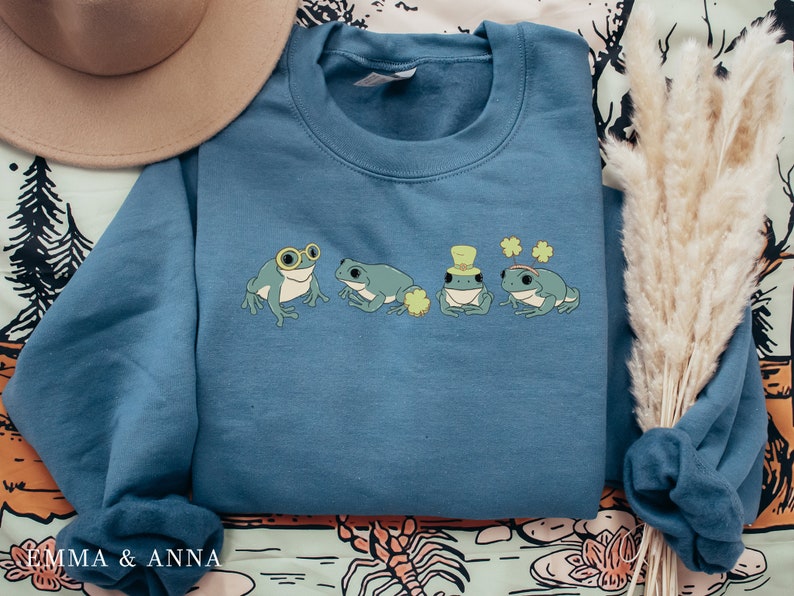 Puede incluir: Una sudadera azul con un gr&aacute;fico de cuatro ranas que llevan sombreros y gafas. Las ranas son verdes y tienen ojos blancos. La sudadera est&aacute; sobre un fondo estampado.