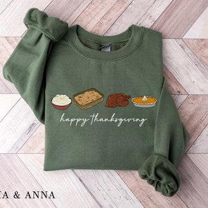 K&ouml;nnte beinhalten: Ein gr&uuml;ner Pullover mit einer wei&szlig;en Grafik eines Truthahns, Kartoffelp&uuml;rees, F&uuml;llungs und K&uuml;rbiskuchens. Der Text "happy Thanksgiving" ist unter dem Essen gedruckt.