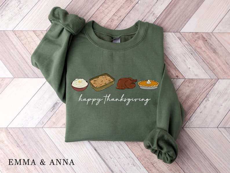 K&ouml;nnte beinhalten: Ein gr&uuml;ner Pullover mit einer wei&szlig;en Grafik eines Truthahns, Kartoffelp&uuml;rees, F&uuml;llungs und K&uuml;rbiskuchens. Der Text "happy Thanksgiving" ist unter dem Essen gedruckt.