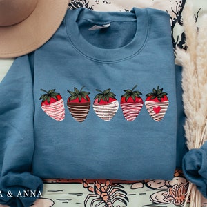 Puede incluir: Una sudadera azul con un gr&aacute;fico de cinco fresas cubiertas de chocolate. Cada fresa tiene una cobertura diferente, incluyendo chocolate blanco, chocolate con chispas y un coraz&oacute;n.