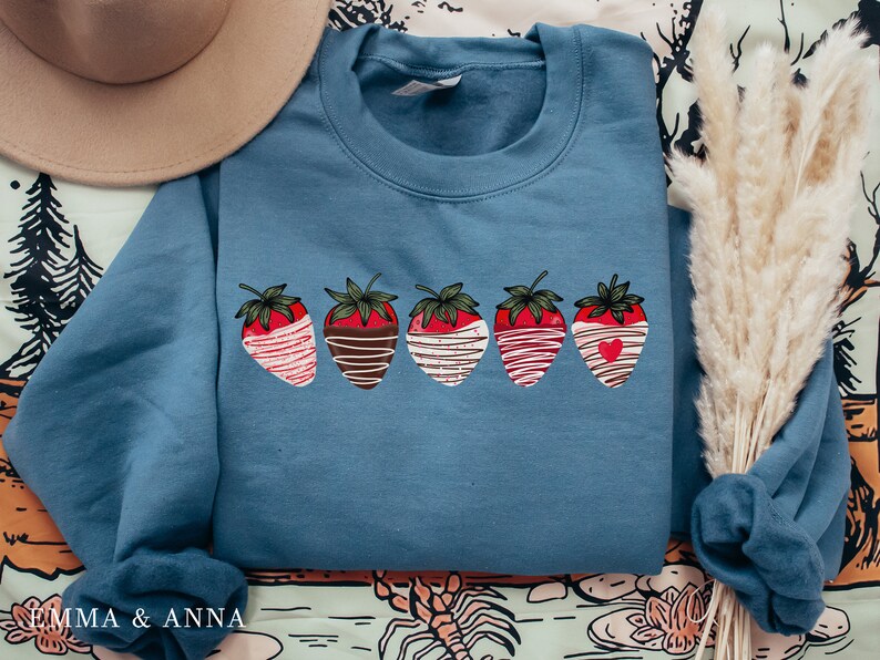 Puede incluir: Una sudadera azul con un gr&aacute;fico de cinco fresas cubiertas de chocolate. Cada fresa tiene una cobertura diferente, incluyendo chocolate blanco, chocolate con chispas y un coraz&oacute;n.