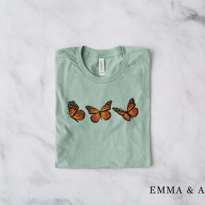 Może przedstawiać: Jasnozielona koszulka z trzema pomarańczowo-czarnymi motylami monarchami. Motyle ułożone są poziomo. Nazwa marki "EMMA & ANNA" jest wydrukowana na dole czarnym kolorem.