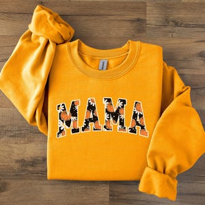 Op de afbeelding: Mosterdgeel sweatshirt met het woord "MAMA" in een zwart-wit bloemen- en pompoenontwerp. De lange mouwen zijn aan de polsen vastgemaakt. Het sweatshirt ligt op een houten oppervlak.
