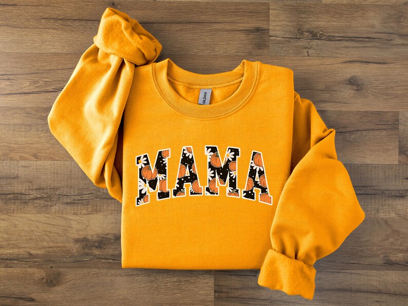 Op de afbeelding: Mosterdgeel sweatshirt met het woord "MAMA" in een zwart-wit bloemen- en pompoenontwerp. De lange mouwen zijn aan de polsen vastgemaakt. Het sweatshirt ligt op een houten oppervlak.