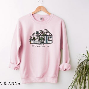 May include: Pale pink sweatshirt featuring a greenhouse illustration with plants inside. The greenhouse is black with green and brown details, and the words "the greenhouse" are below. The brand name "EMMA & ANNA" is at the bottom.