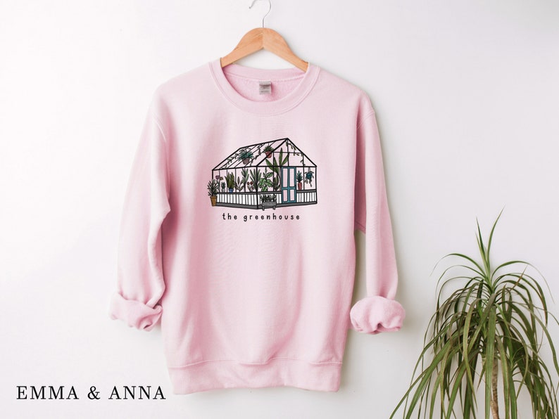 May include: Pale pink sweatshirt featuring a greenhouse illustration with plants inside. The greenhouse is black with green and brown details, and the words "the greenhouse" are below. The brand name "EMMA & ANNA" is at the bottom.