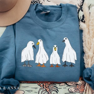 May include: A blue sweatshirt with a graphic of four white ghost ducks. The ducks are lined up in a row and have different expressions on their faces.