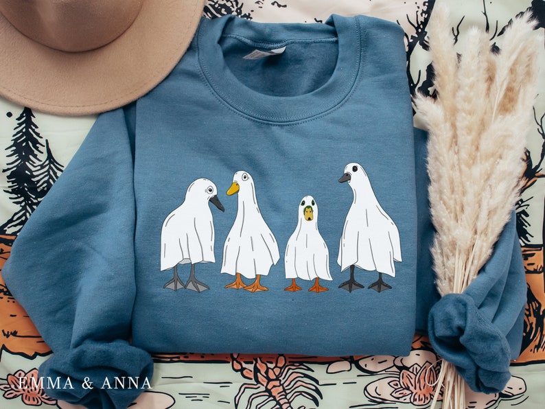 May include: A blue sweatshirt with a graphic of four white ghost ducks. The ducks are lined up in a row and have different expressions on their faces.